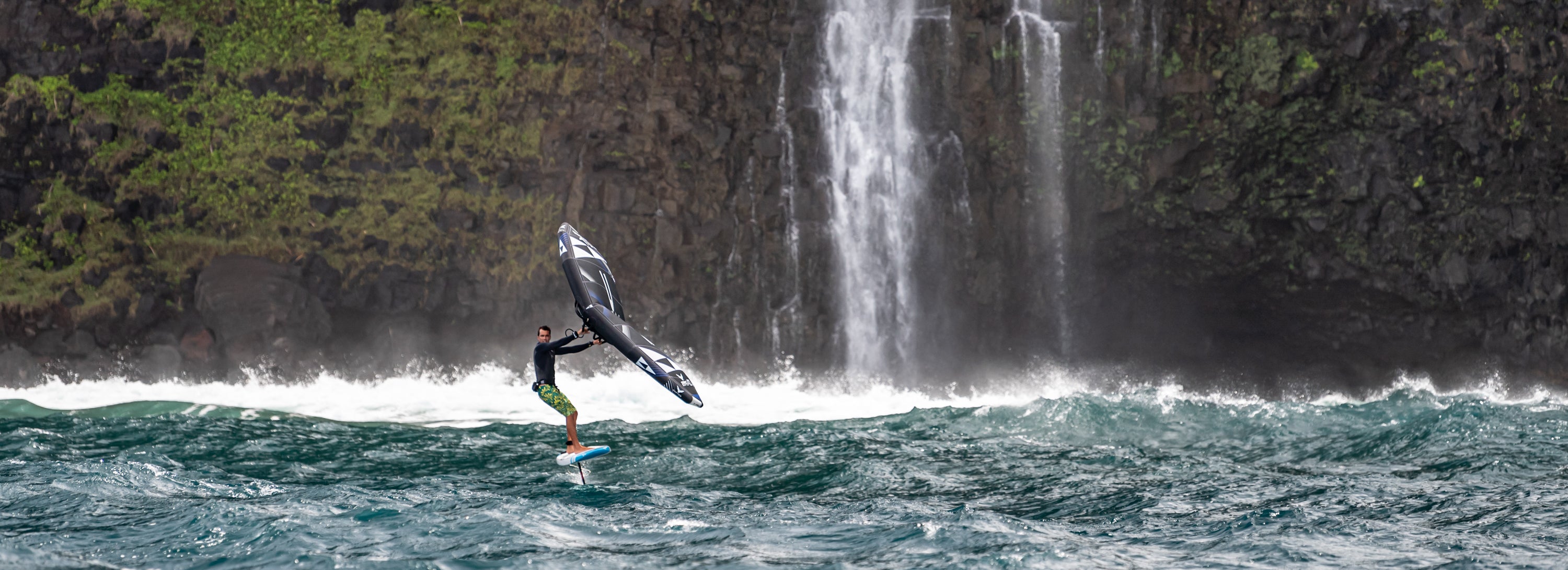 Wingsurfer riding a blue and white raptor foil board with a black wing and a slash foil in front of a massive tropical waterfall and dark volcanic cliffs - SIC MAUI