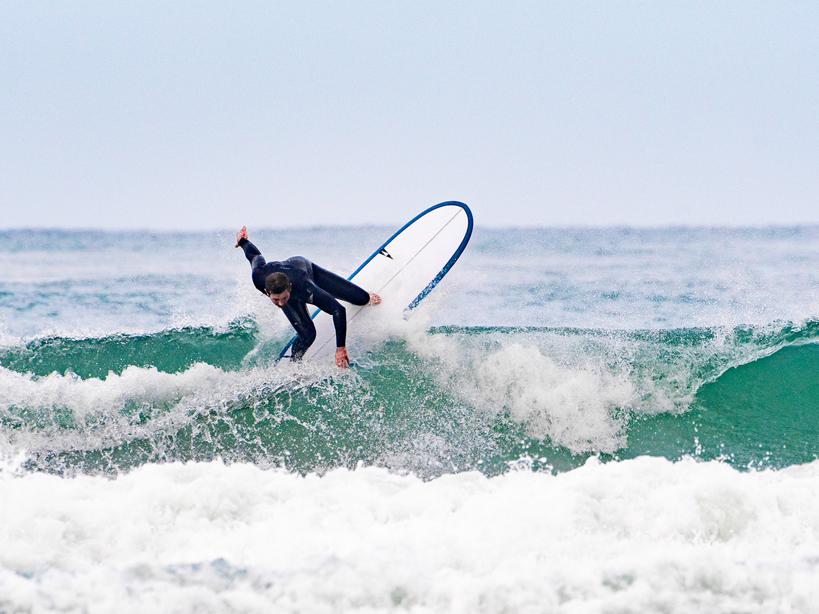 A surfer in a dark wetsuit carves a sharp turn on a wave, spray flying, riding a Pick Pocket surfboard with blue trim against an overcast sky SIC MAUI