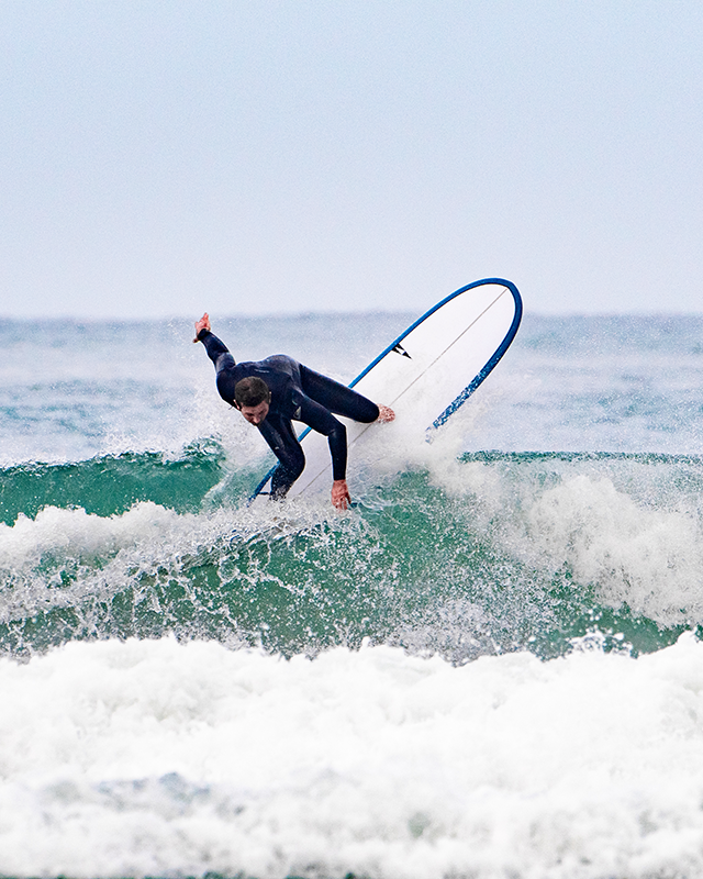 A surfer in a dark wetsuit carves a sharp turn on a wave, spray flying, riding a Pick Pocket surfboard with blue trim against an overcast sky SIC MAUI