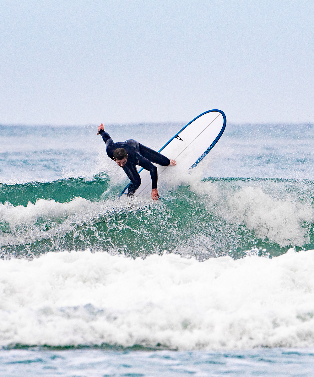 A surfer in a dark wetsuit carves a sharp turn on a wave, spray flying, riding a Pick Pocket surfboard with blue trim against an overcast sky SIC MAUI