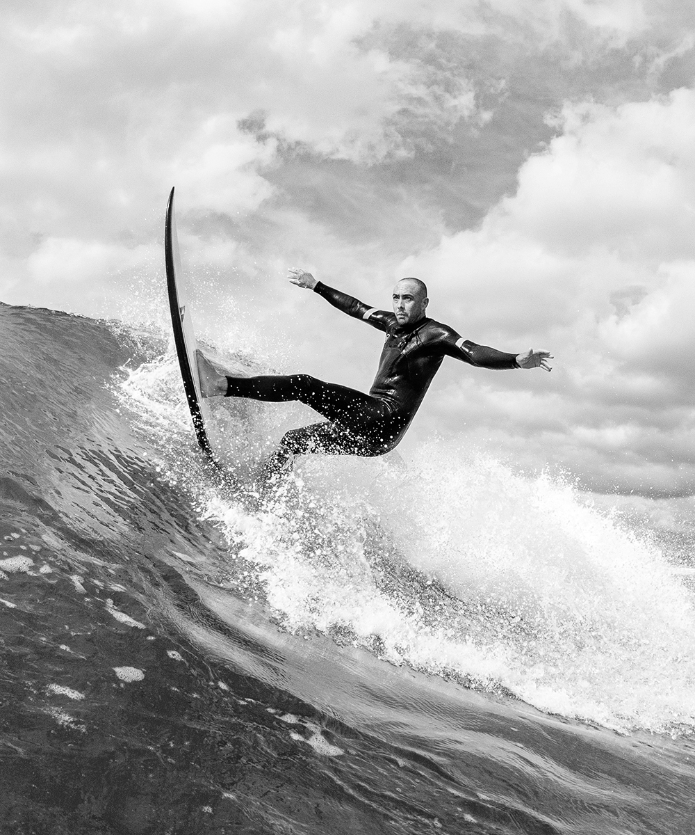 Black and white photo of a man in a wetsuit performing a vertical snap with a SIC Maui shortboard, throwing spray.