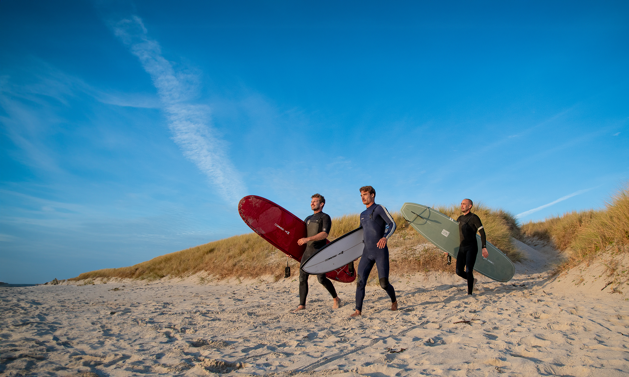 Three surfers in full wetsuits walk along a sandy beach near dunes, carrying their SIC Maui surfboards (maroon, white, and olive green) under a clear blue sky.
