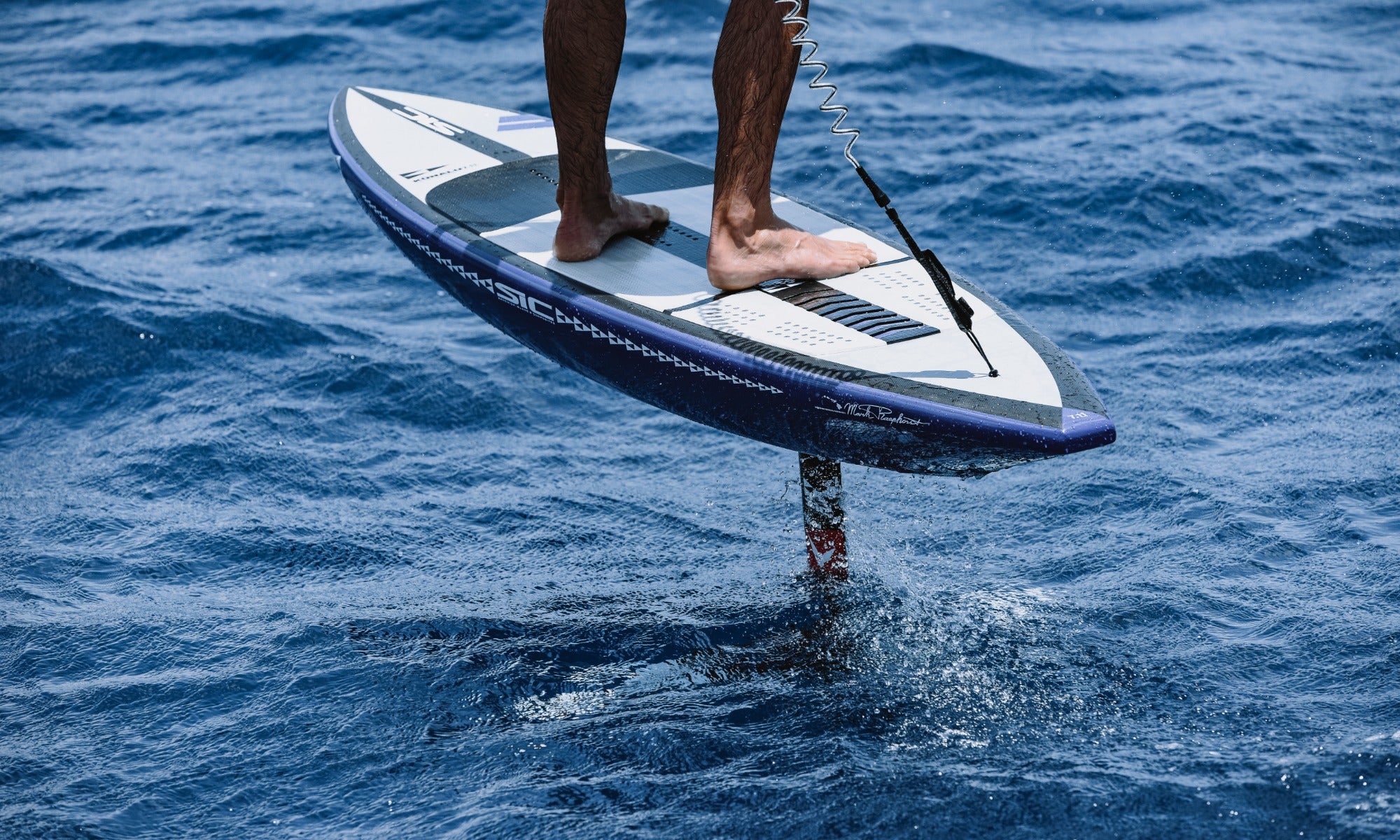 Close-up view of bare feet on a SIC Maui foil board with coiled leash in the blue ocean.