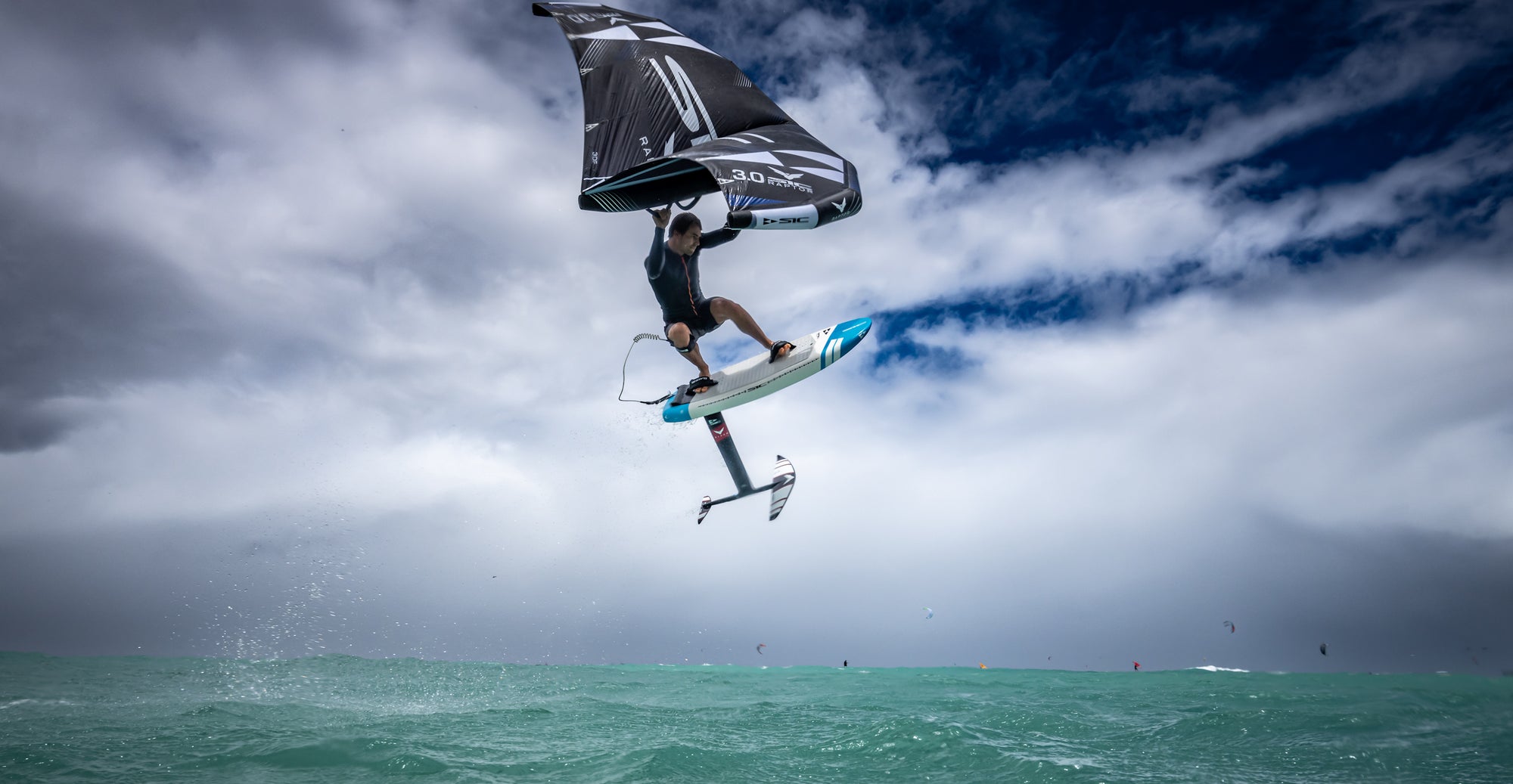 A wingsurfer soaring high above the teal water, holding a black 3.0 Raptor wing, on a white and blue raptor foil board with a slash foil, under a dramatic, cloudy sky - SIC MAUI
