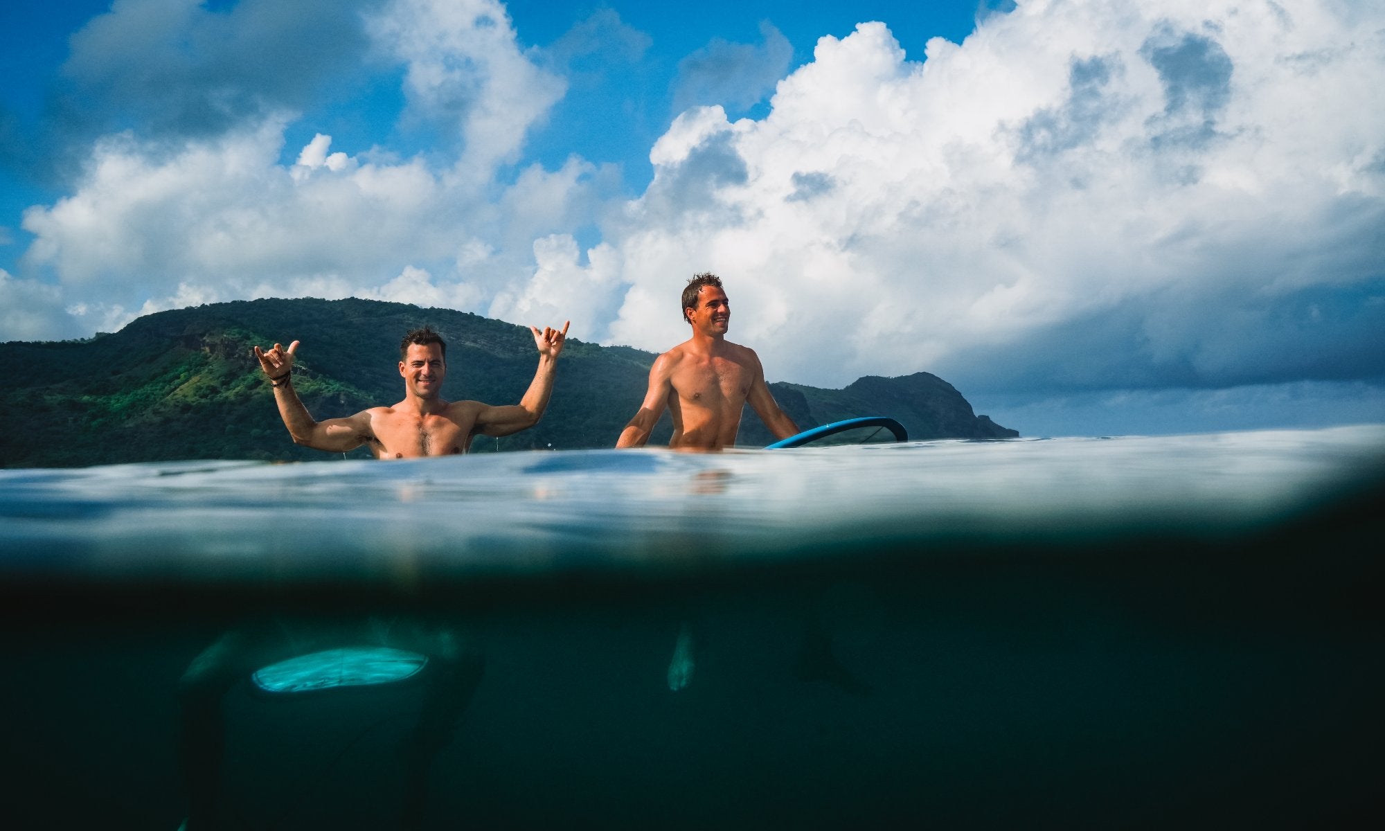 Two surfers, Ludovic Teulade and is brother, sitting on their SIC Maui surfboards in water, one making a shaka sign against a backdrop of lush green mountains.