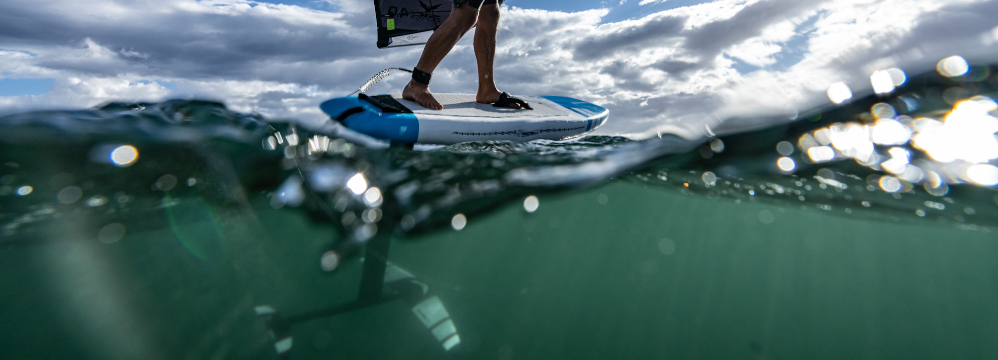 Close-up split shot of a rider on a blue and white SIC Maui wing foil board with the Slash foil, showing the hydrofoil submerged in green water under a cloudy sky.