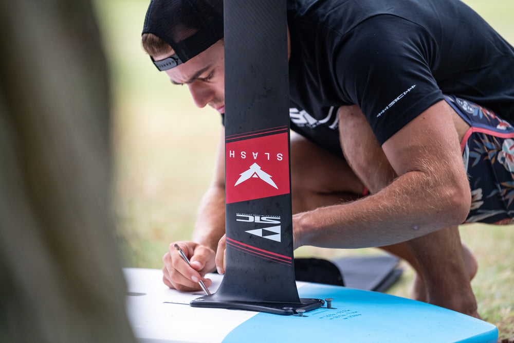 Close-up of a rider assembling a black SIC Maui Slash foil mast onto a blue and white raptor board using a tool, focusing on the mounting hardware.