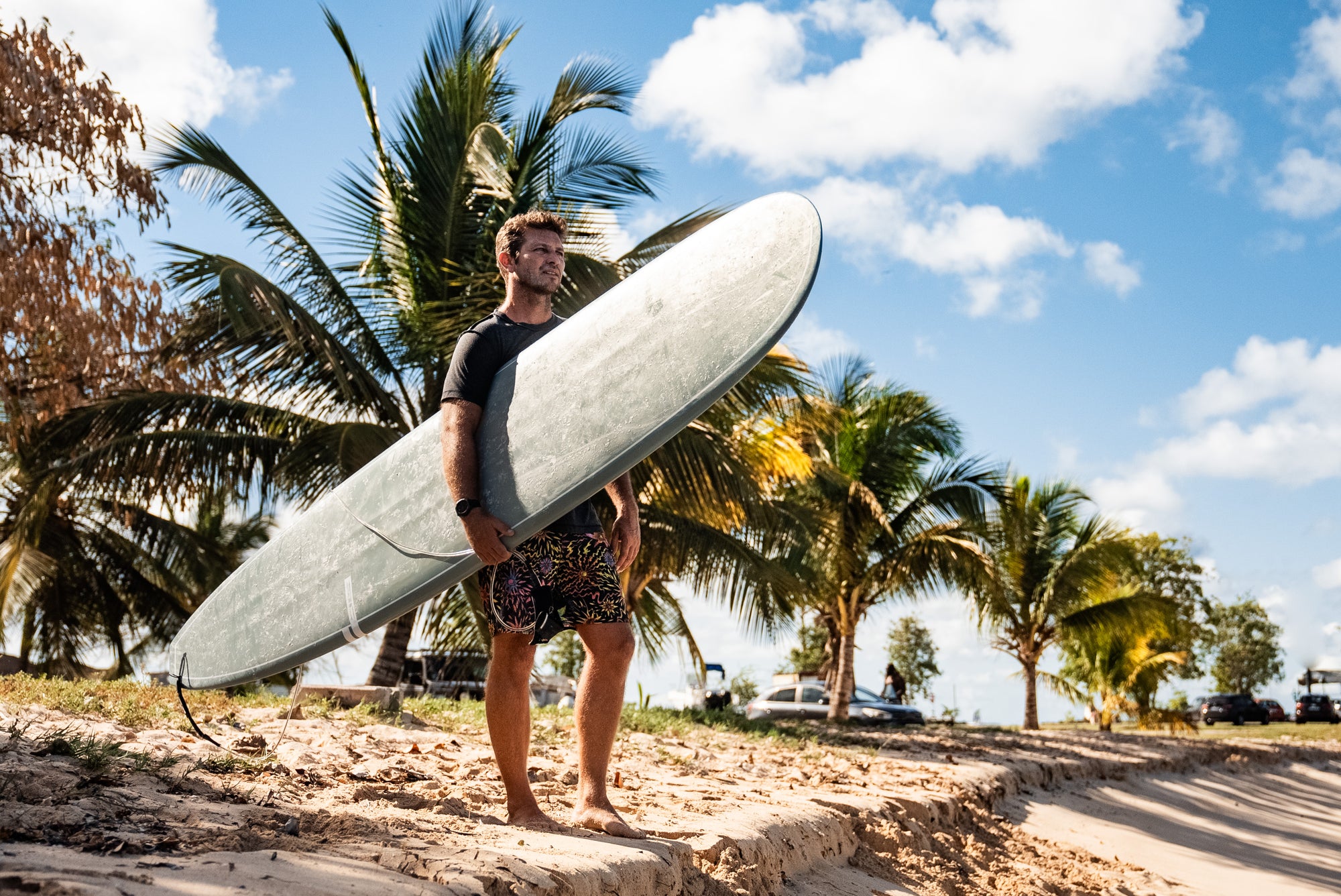 Man standing on a sandy beach holding a green Smuggler longboard surfboard under one arm, looking up, with palm trees and a cloudy sky behind him - SIC MAUI