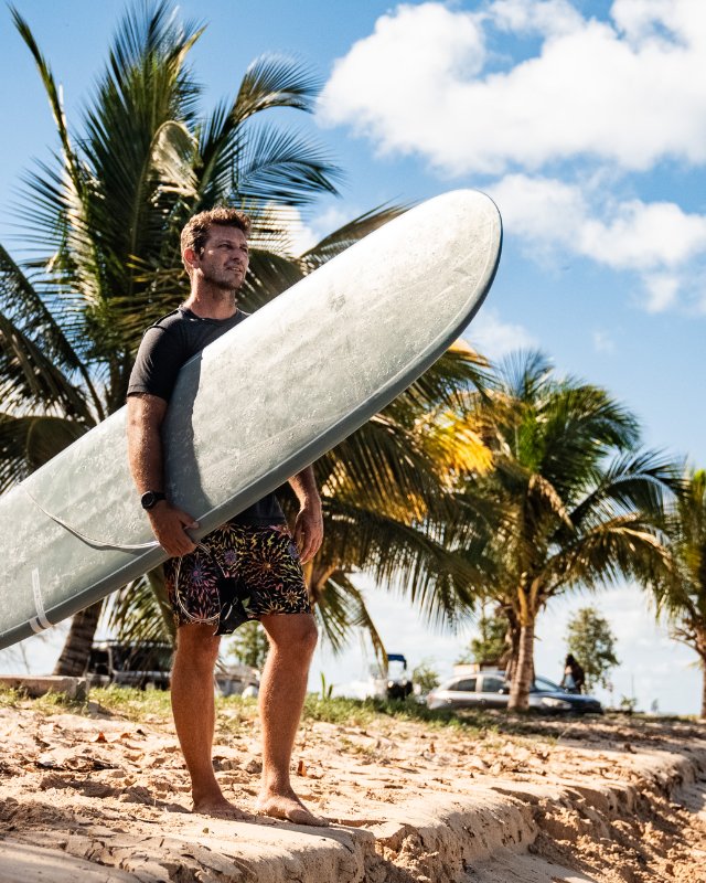 Man standing on a sandy beach holding a green Smuggler longboard surfboard under one arm, looking up, with palm trees and a cloudy sky behind him - SIC MAUI