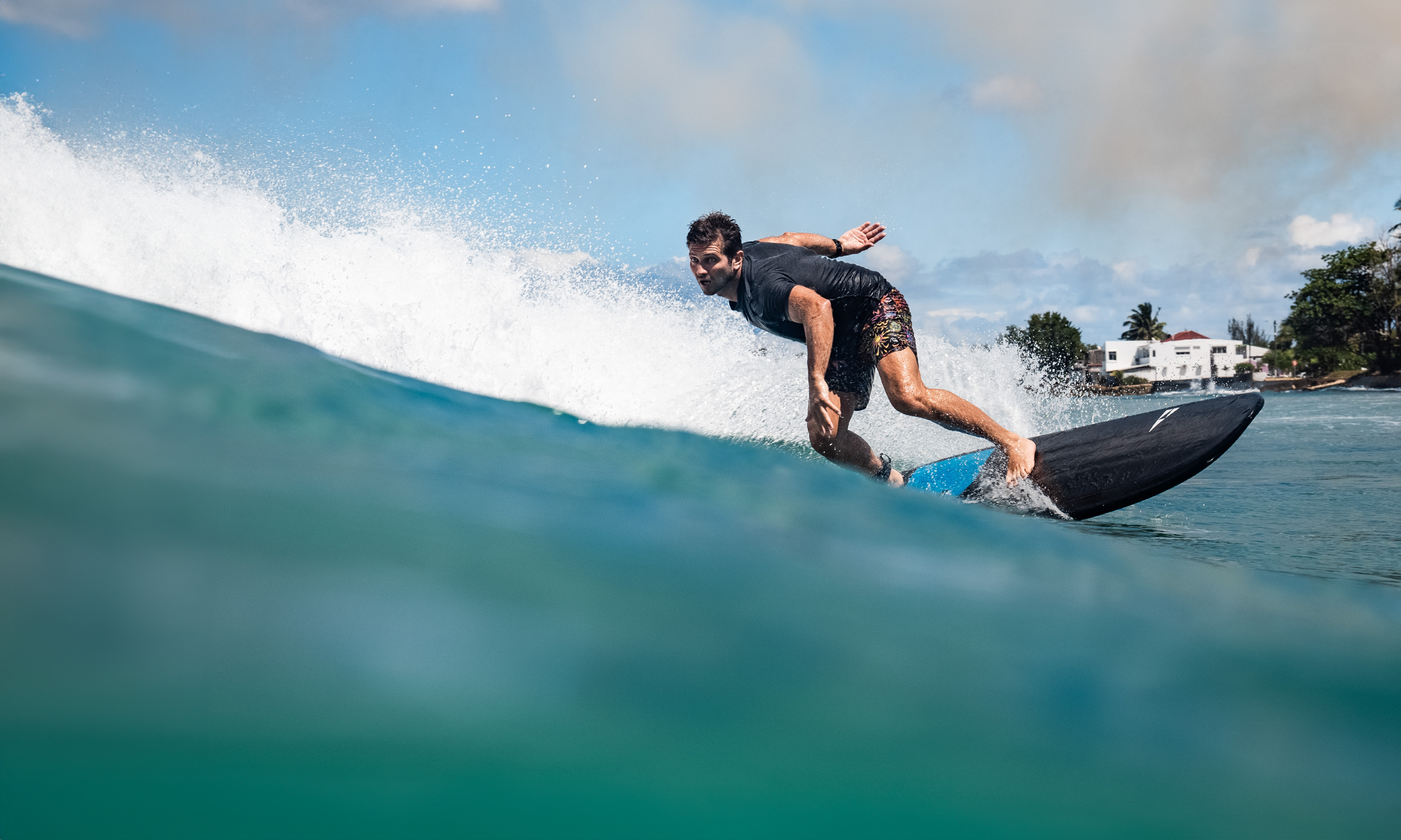 Surfer carving a wave on a black and blue Phantom surfboard with white spray and tropical coastline in the background, high-performance action, SIC MAUI.