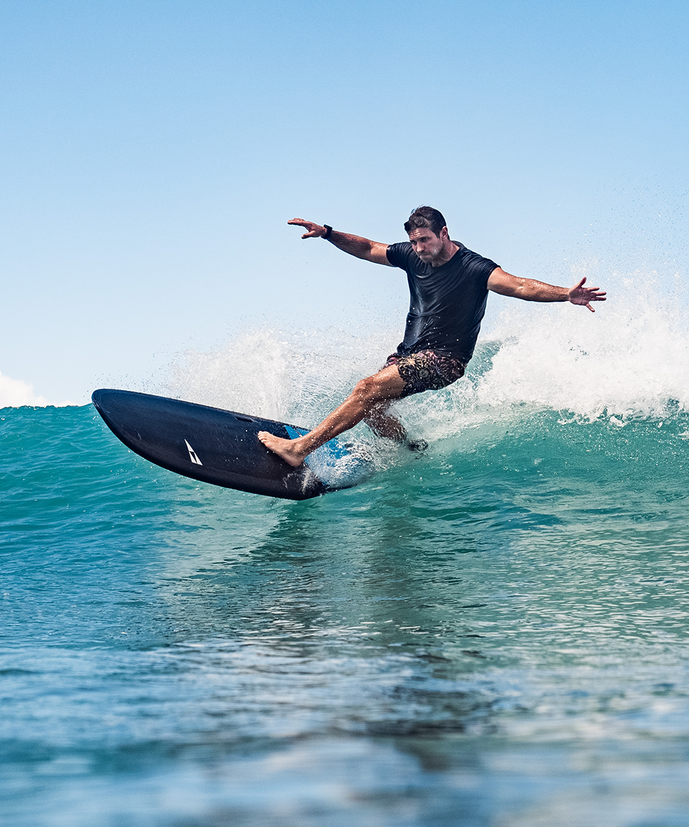 Surfer performing a dynamic turn on a black Phantom surfboard, creating white water spray against a clear blue sky and turquoise ocean - SIC MAUI