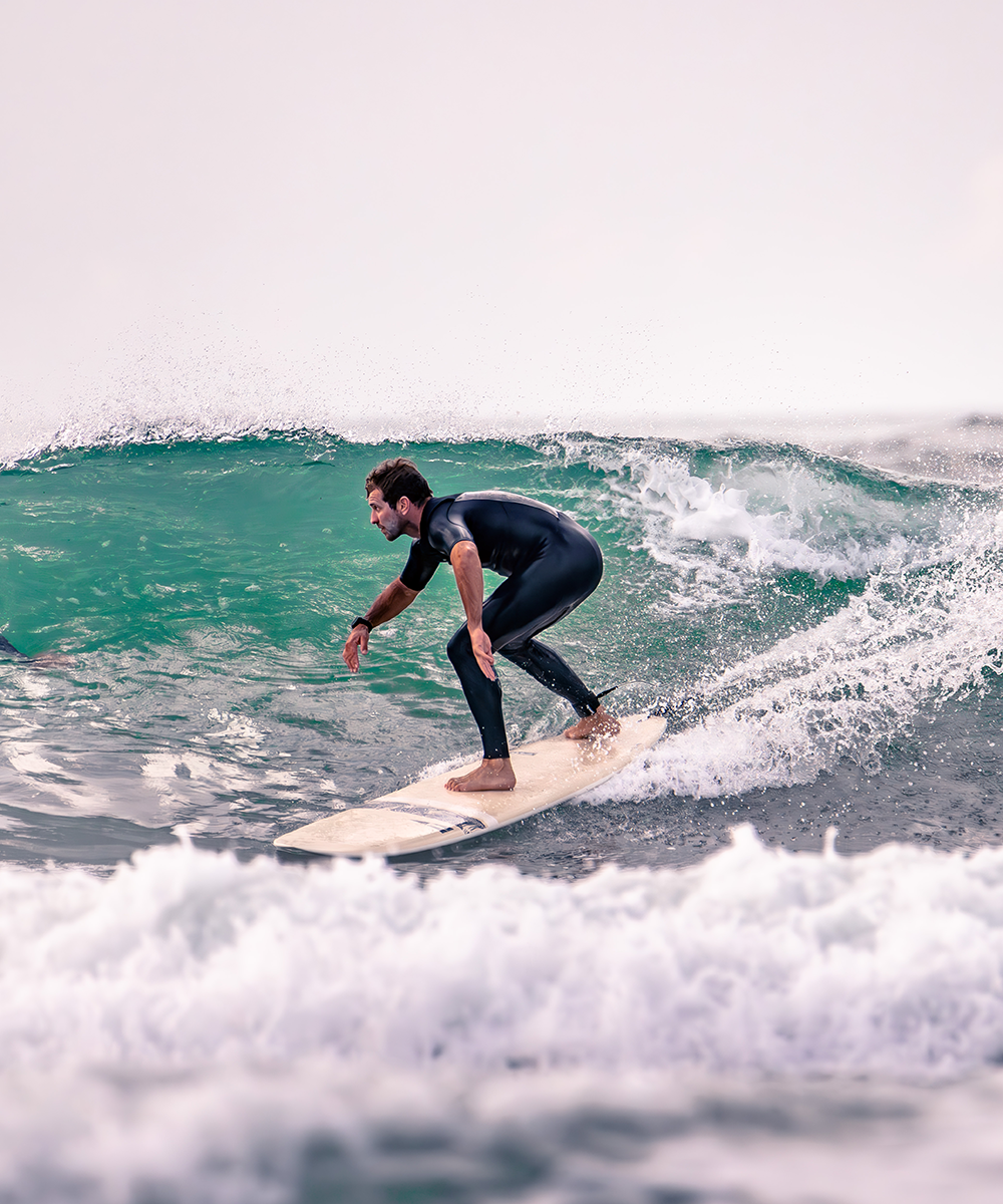 Surfer in a black wetsuit riding a turquoise wave on a drifter surfboard, showcasing balance and performance in a bright ocean setting - SIC MAUI