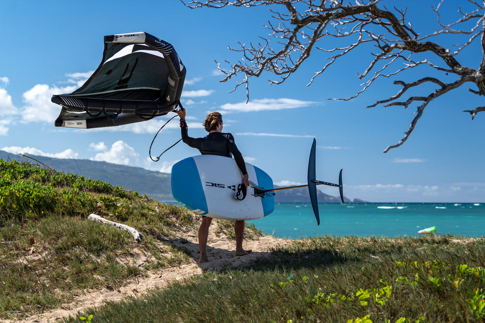 Rider walking toward a turquoise bay holding a black SIC Maui wing and a blue and white raptor foil board, with tropical hills and a blue sky in the background.