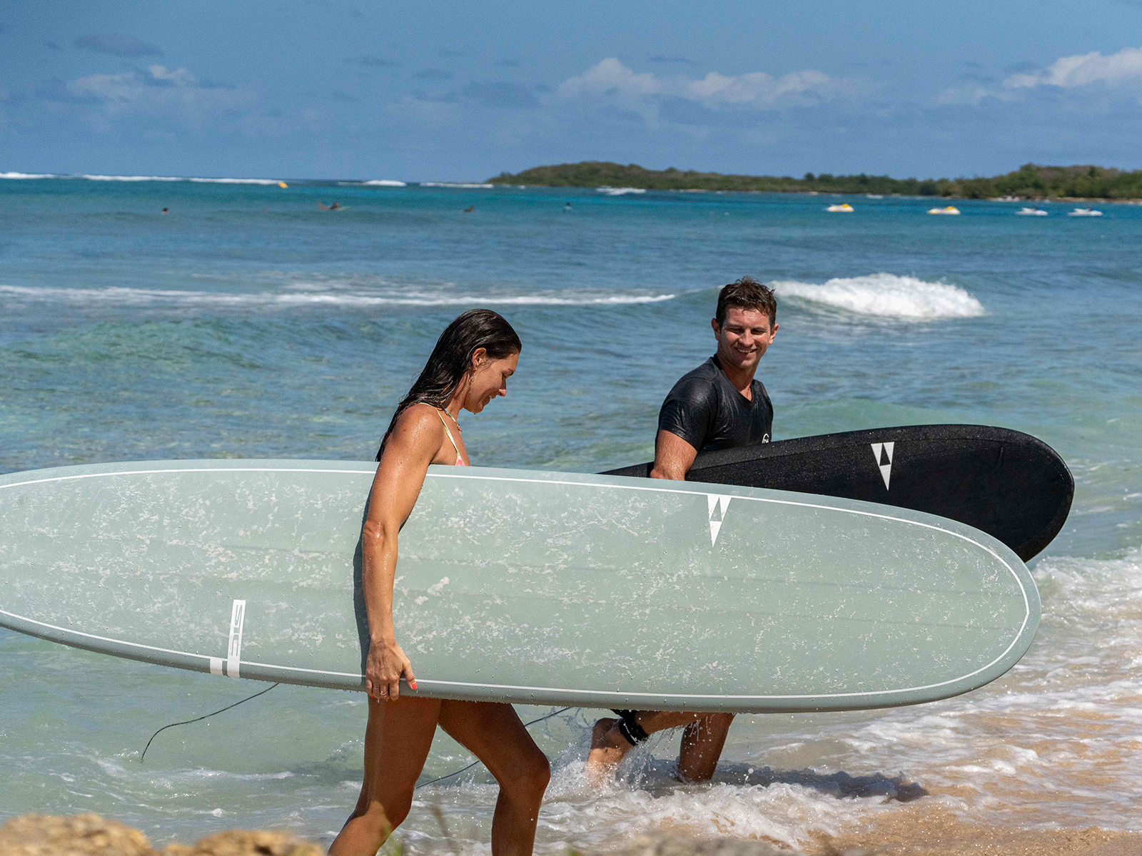 Two surfers walk out of the tropical water onto the sand, carrying a sage green Smuggler Longboard and a black Phantom surfboard under a bright, sunny blue sky SIC MAUI