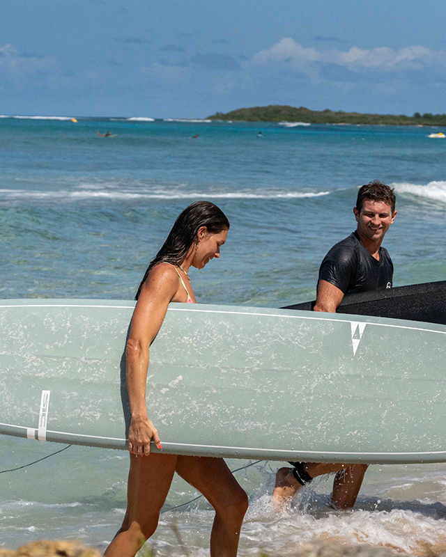 Two surfers walk out of the tropical water onto the sand, carrying a sage green Smuggler Longboard and a black Phantom surfboard under a bright, sunny blue sky SIC MAUI